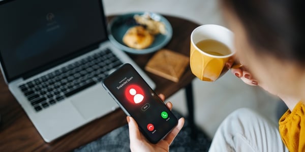 Over the shoulder view of young Asian woman receiving an incoming suspected call from unknown caller on her smartphone and rejecting the call at home. Device screen showing warning sign as detected by the network provider. Phone scam and fraud concept
