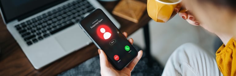 Over the shoulder view of young Asian woman receiving an incoming suspected call from unknown caller on her smartphone and rejecting the call at home. Device screen showing warning sign as detected by the network provider. Phone scam and fraud concept